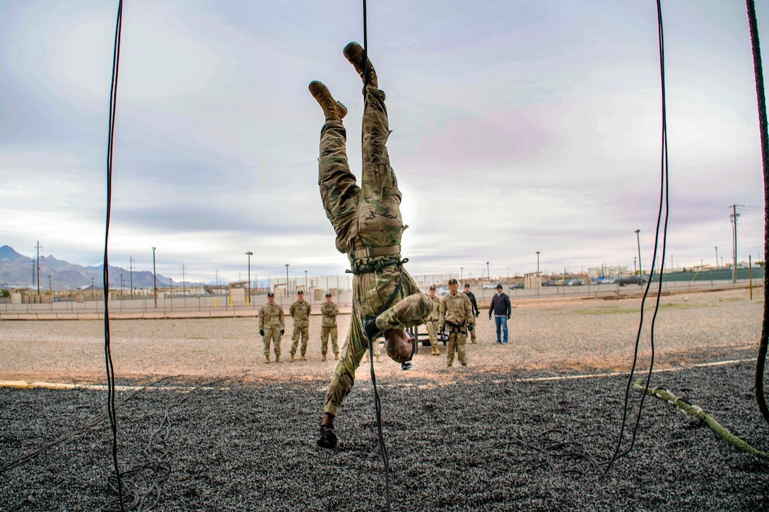 An air assault instructor reaches out to touch the ground beneath his head while hanging upside down.