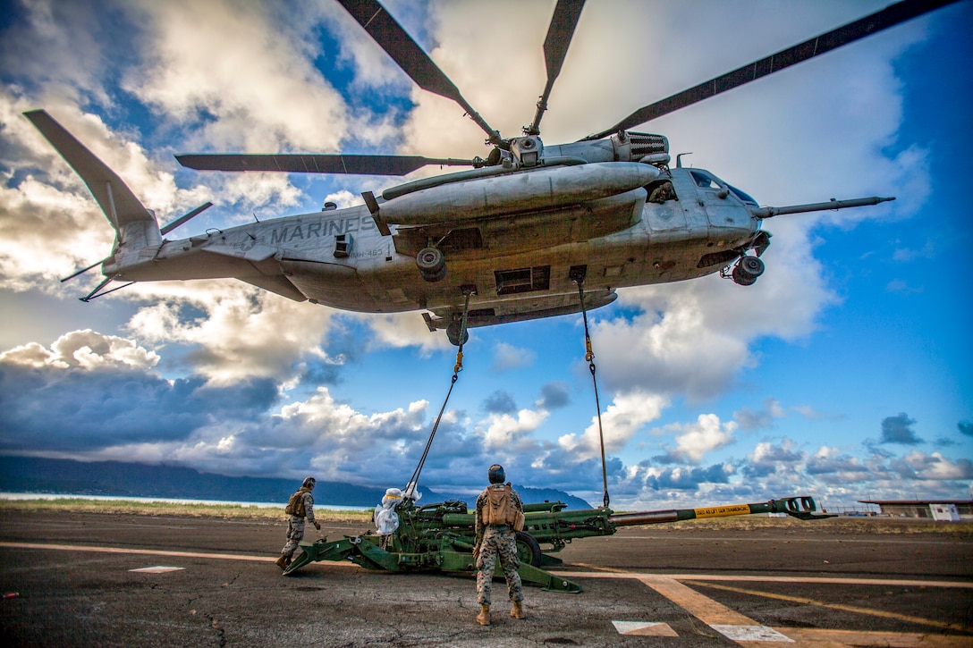 A helicopter attached to a howitzer takes off as Marines watch.