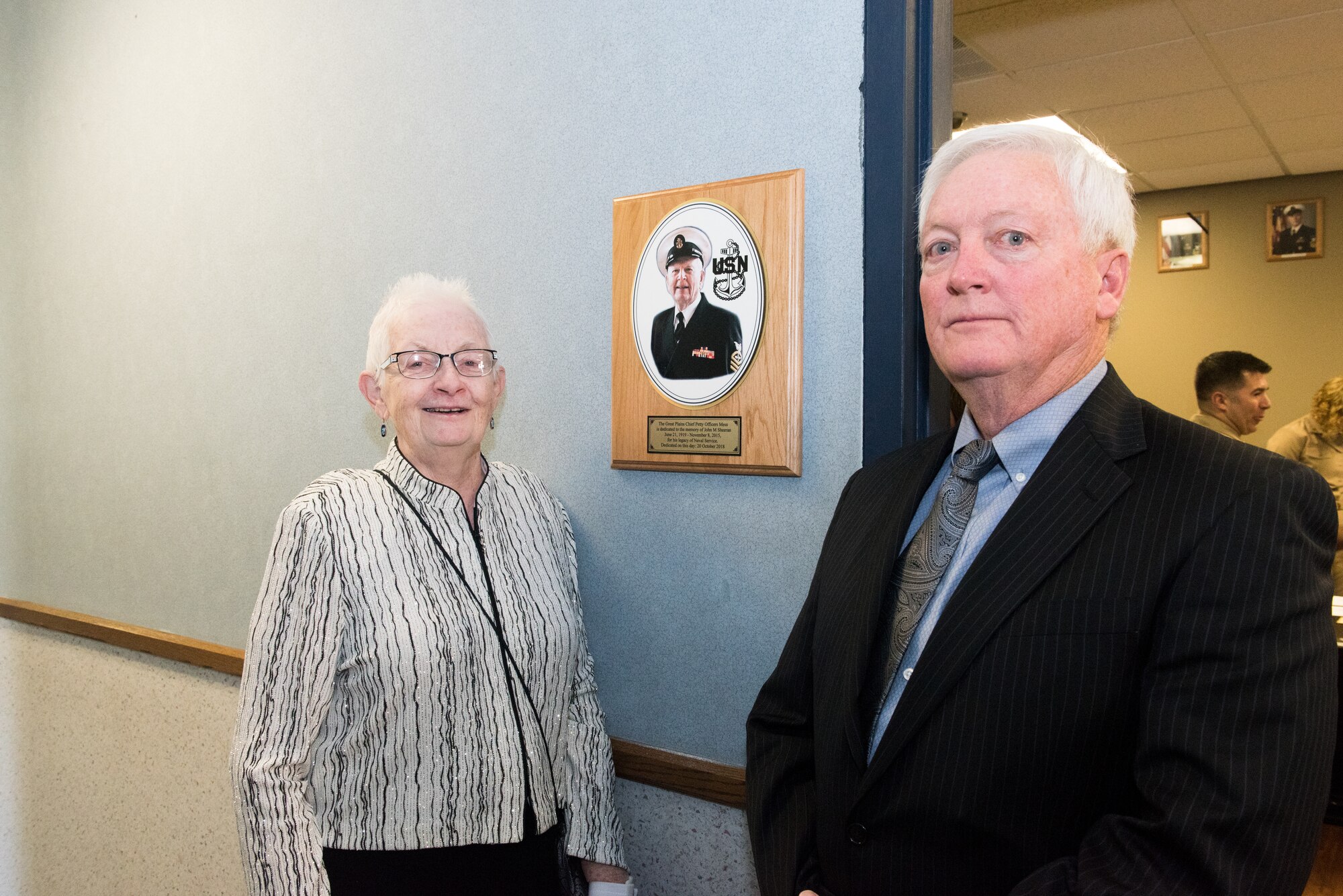 Linda Sheeran, wife of U.S. Navy veteran and retired Chief Petty Officer John Sheeran, and his son, John, pose in front of the plaque outside the Great Plains Chief Petty Officer Association Chief’s Mess that names the room after John following a ceremony at the 55th Operations Support Squadron Oct. 20, 2018.