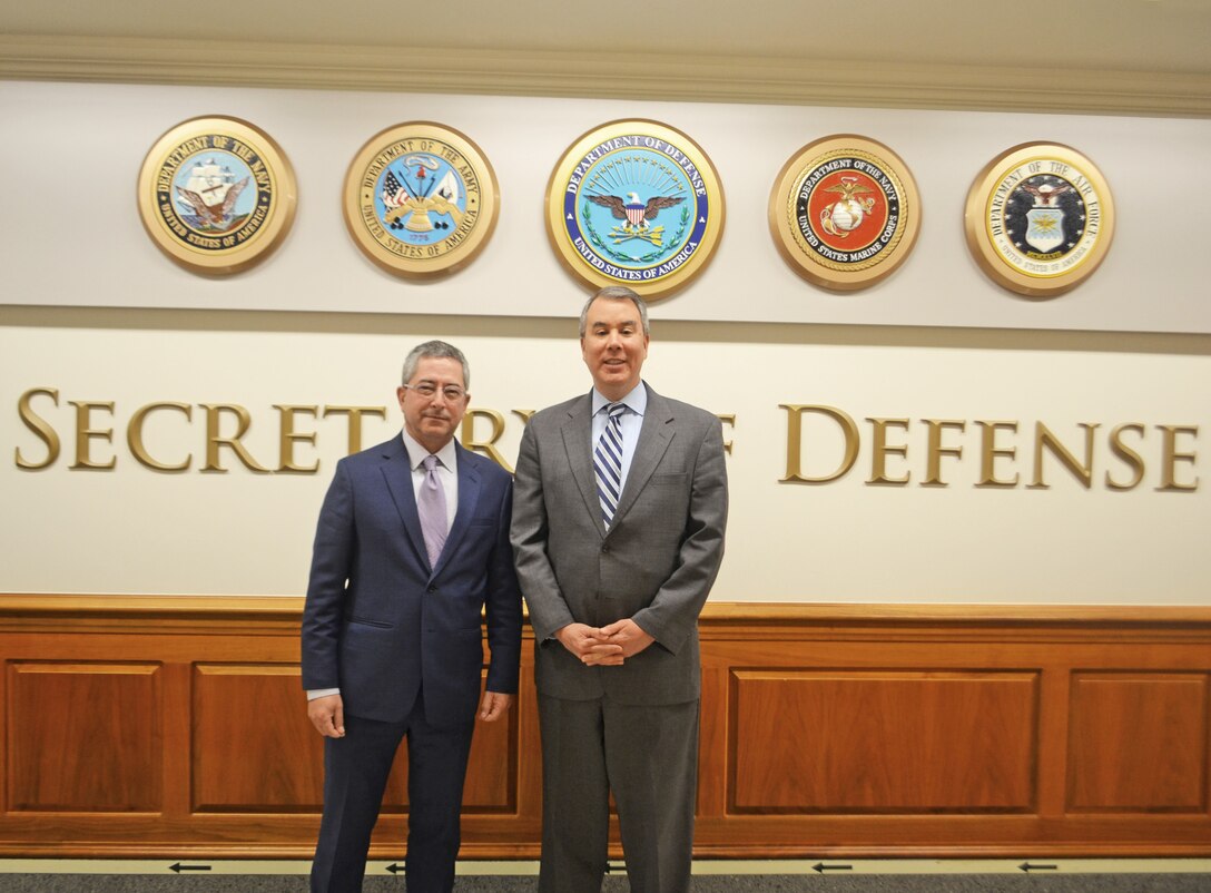 Two men in suits pose for a photo with military seals in the background.