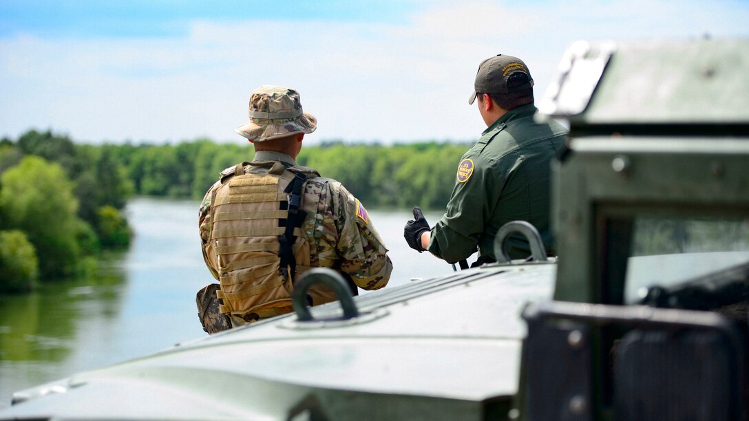 A service member and a border patrol agent, shown from behind, talk while standing by a river.