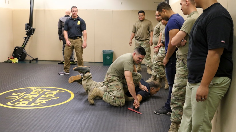 A guardsman wrestles with an instructor in a gym as other troops watch.