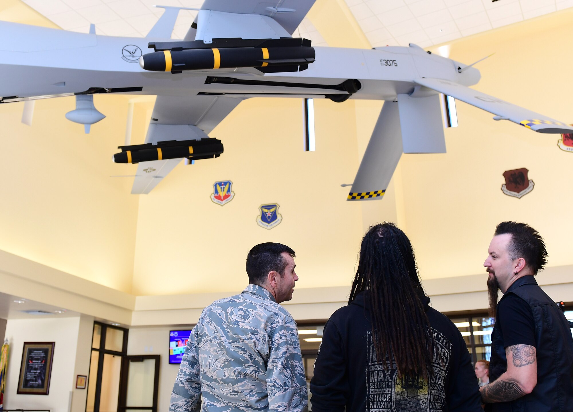 Col. James Price, 432nd Wing/432nd Air Expeditionary Wing vice commander, John Moyer, Disturbed bassist,  and Mike Wengren, Disturbed drummer, observe an MQ-1 Predator model at Creech Air Force Base, Nevada, Oct. 23, 2018. Wing leadership greeted the band members before Disturbed and the USO team were walked through a mission brief. (U.S. Air Force photo by Senior Airman Christian Clausen)