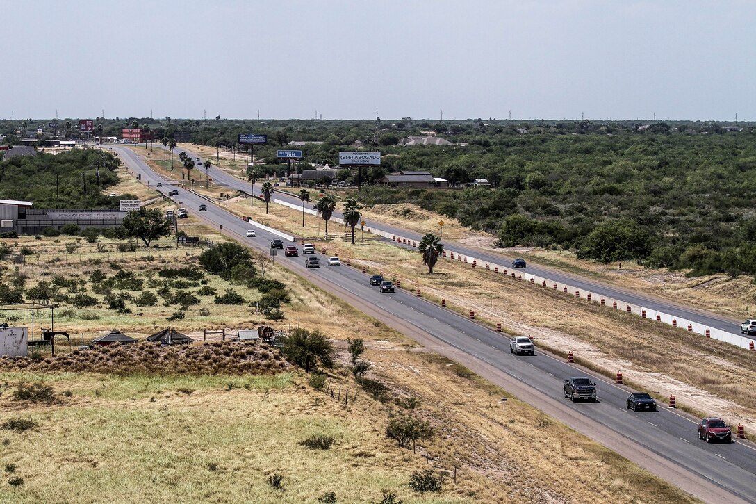 An oblique aerial view of two strips of highway over mostly flat, green terrain.