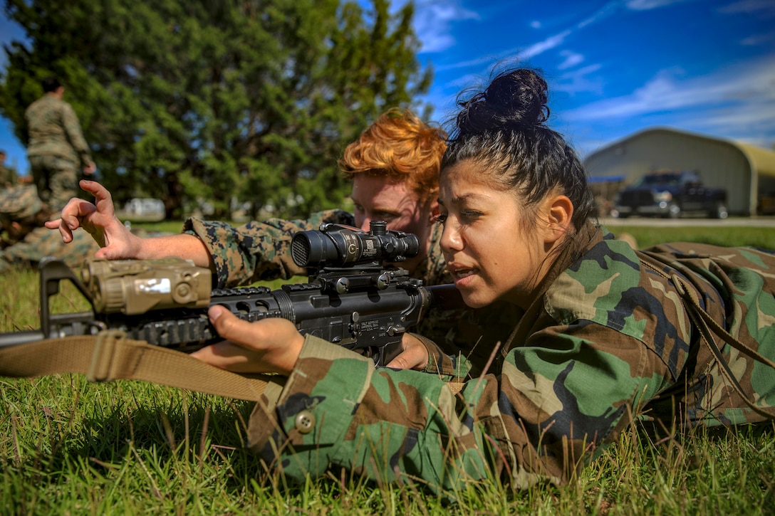 A woman points a rifle while lying in a field as a Marine provides direction beside her.