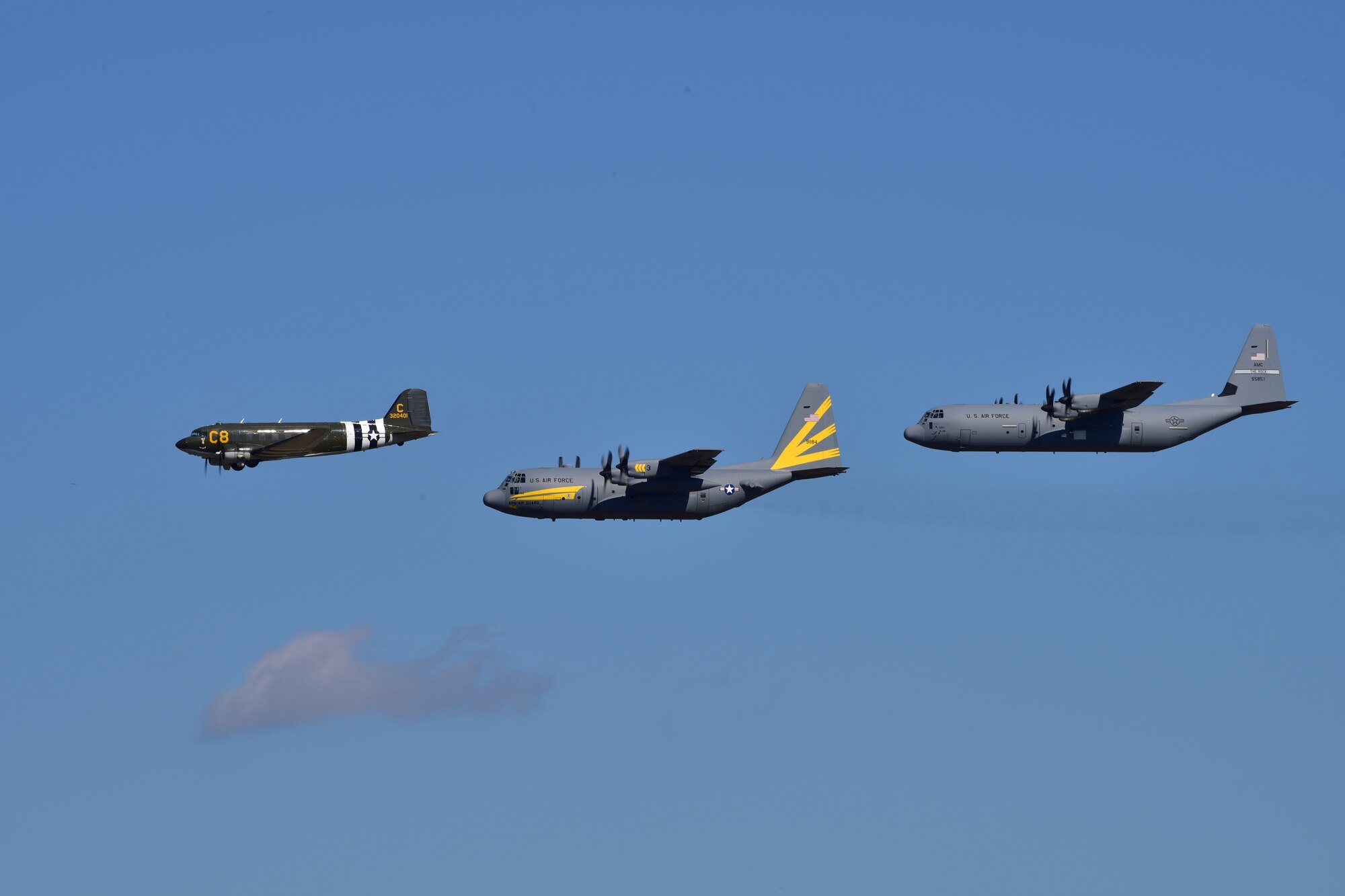 A green Douglas C-47 Skytrain aircraft, a gray C-130H aircraft with yellow stripes, and a gray C-130J aircraft fly in the sky