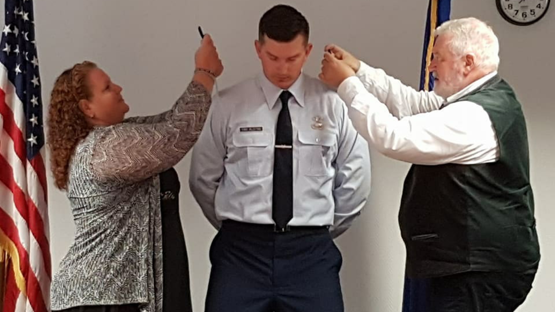 Man in uniform stands with head bowed as a woman to his left and a man to his right make adjustments to his uniform.