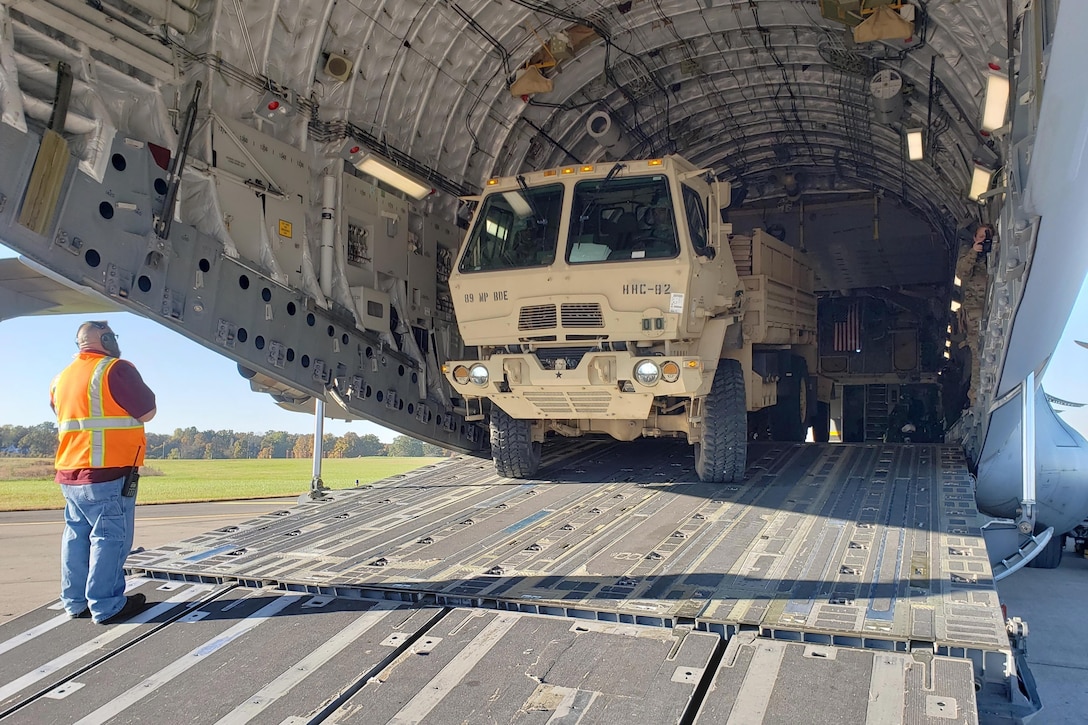 A military vehicle backs into an open aircraft as a man in an orange vest watches.
