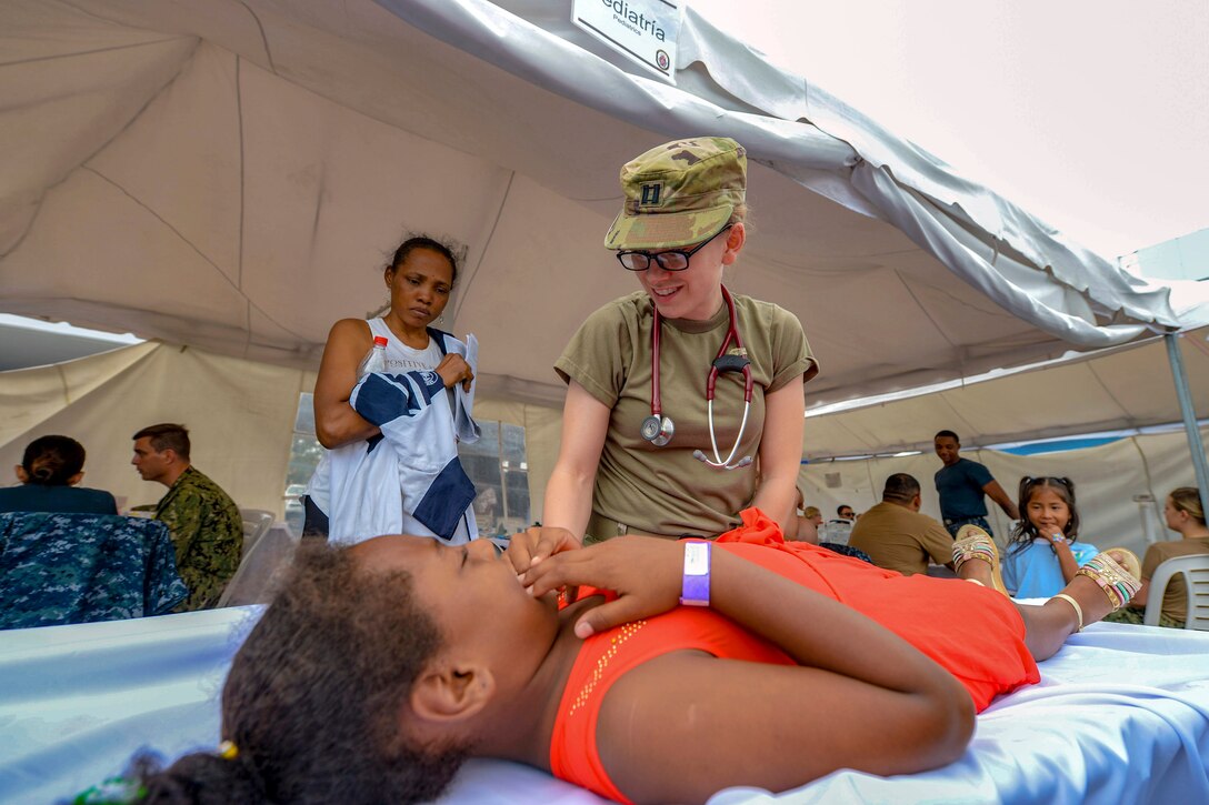 A doctor smiles while talking to a girl lying on a cot by a tent outside.