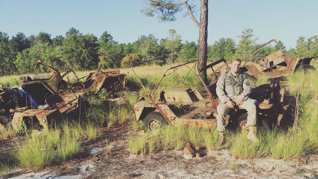 A soldier sits on a vehicle in tall grass.