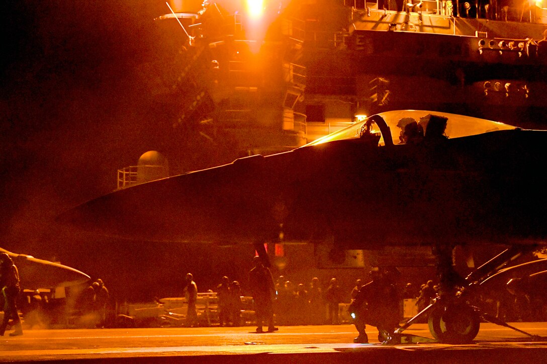 Service members work around a jet on a flight deck illuminated by orange light at night.