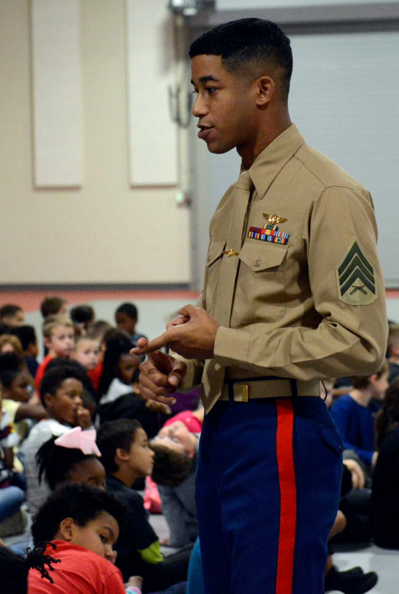 A young black male in Marine Corps tan shirt and blue pants stands in the middle of a group of children as he talks to them.