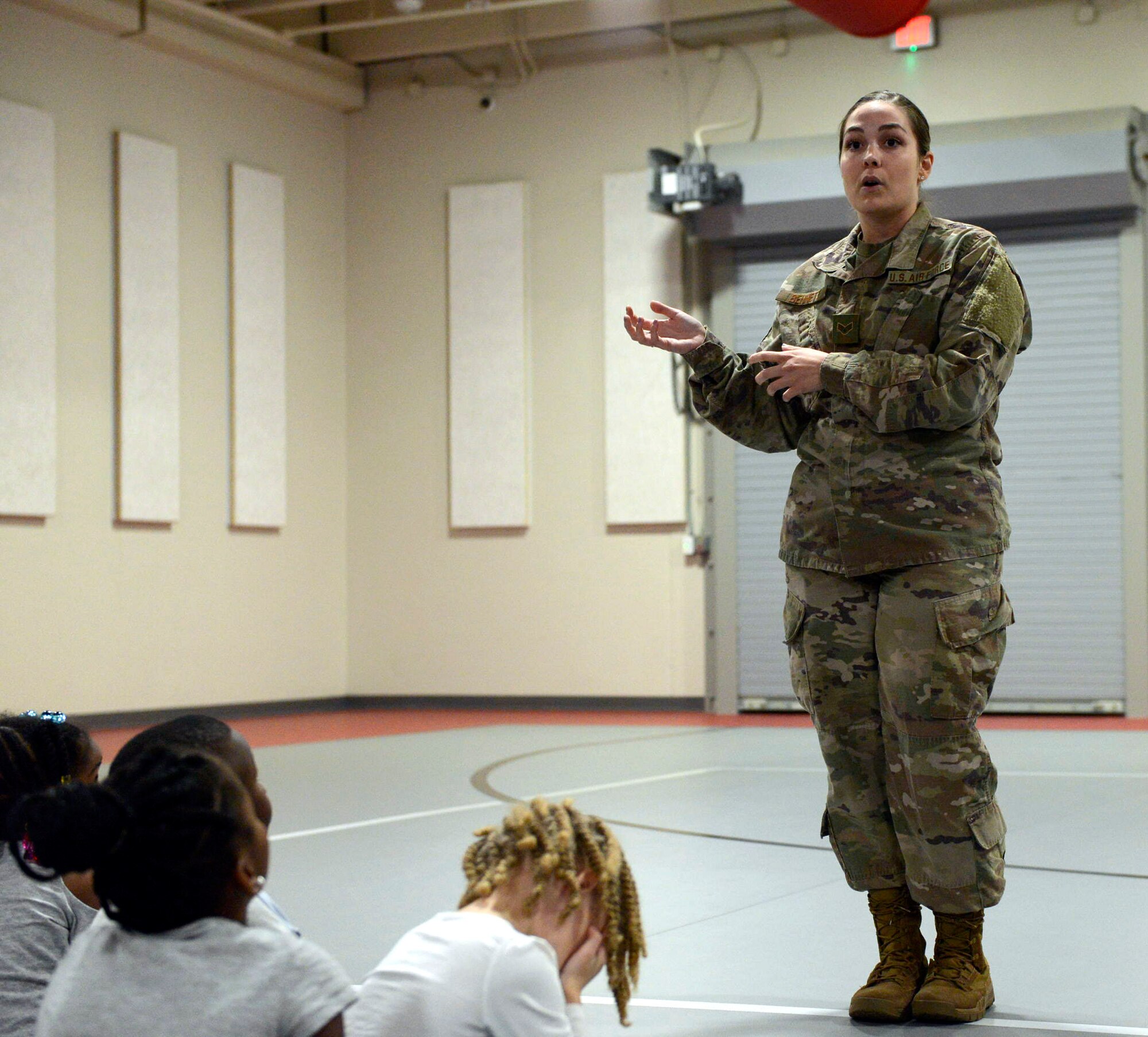 A female in camouflage talks to students in a gym.