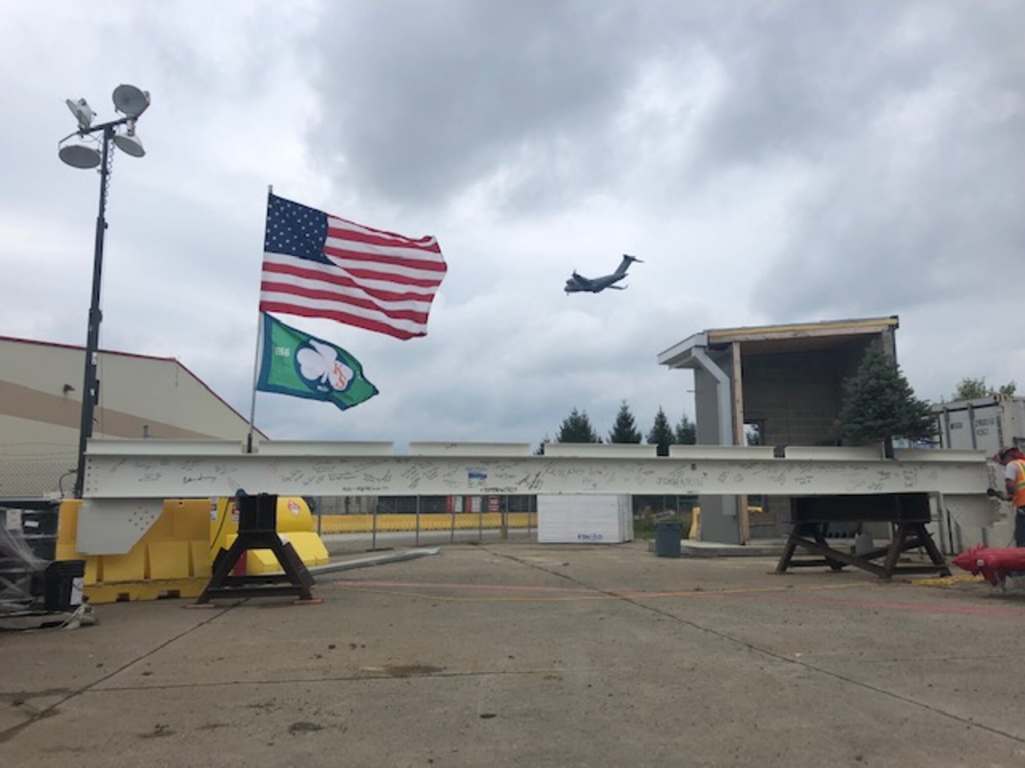 A C-17 aircraft assigned to the 911th Airlift Wing flies over the 911th AW as a signed beam ready to be installed in the hangar which will house the aircraft sits at the Pittsburgh International Airport Air Reserve Station, Pennsylvania, Oct. 4, 2017. This beam, signed by base members, will be the last to be installed in the roof of the hangar.(U.S. Air Force courtesy photo by Master Sgt. Frank Powell)