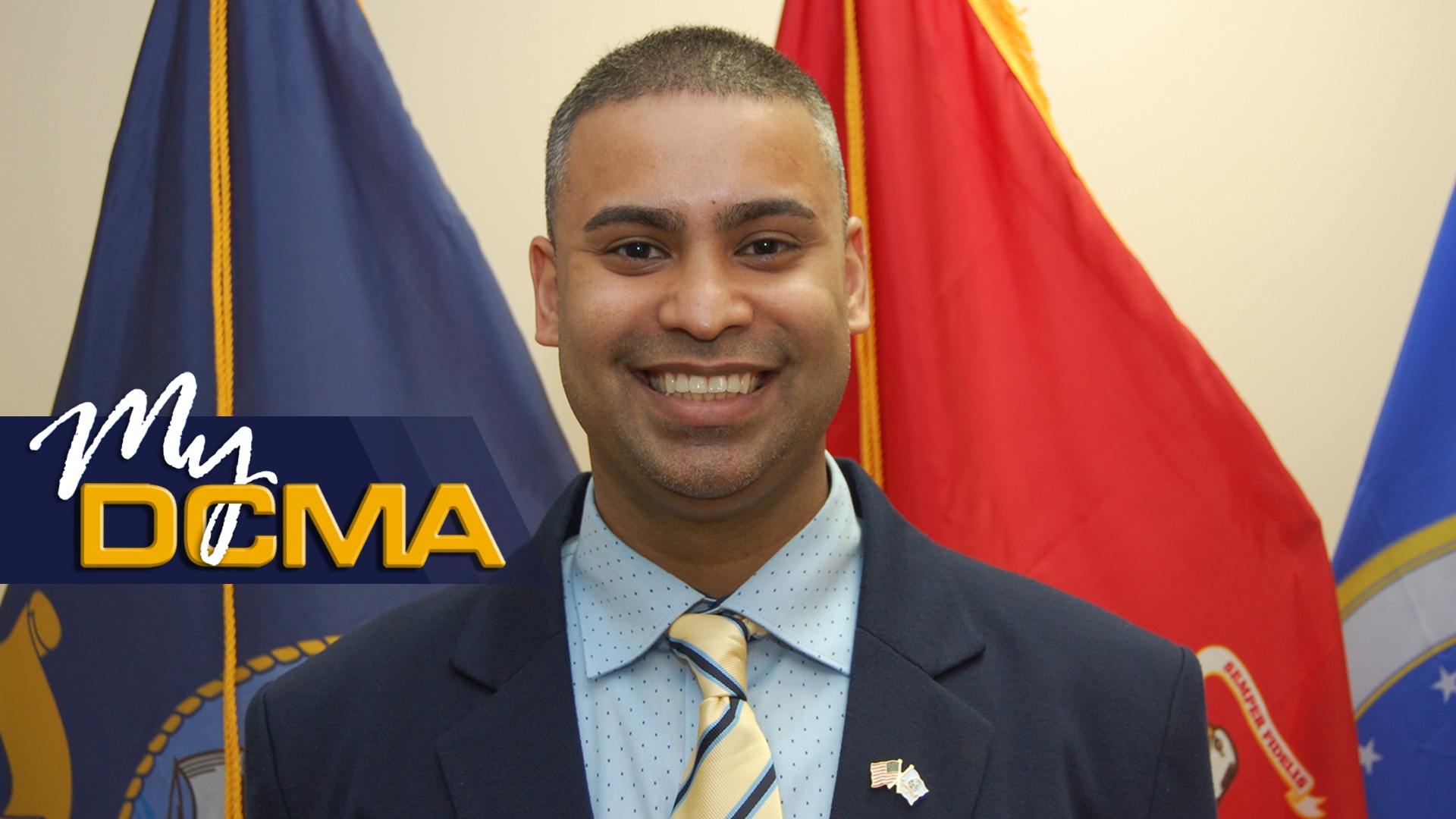A smiling man in a suit stands in front of the various DCMA flags.