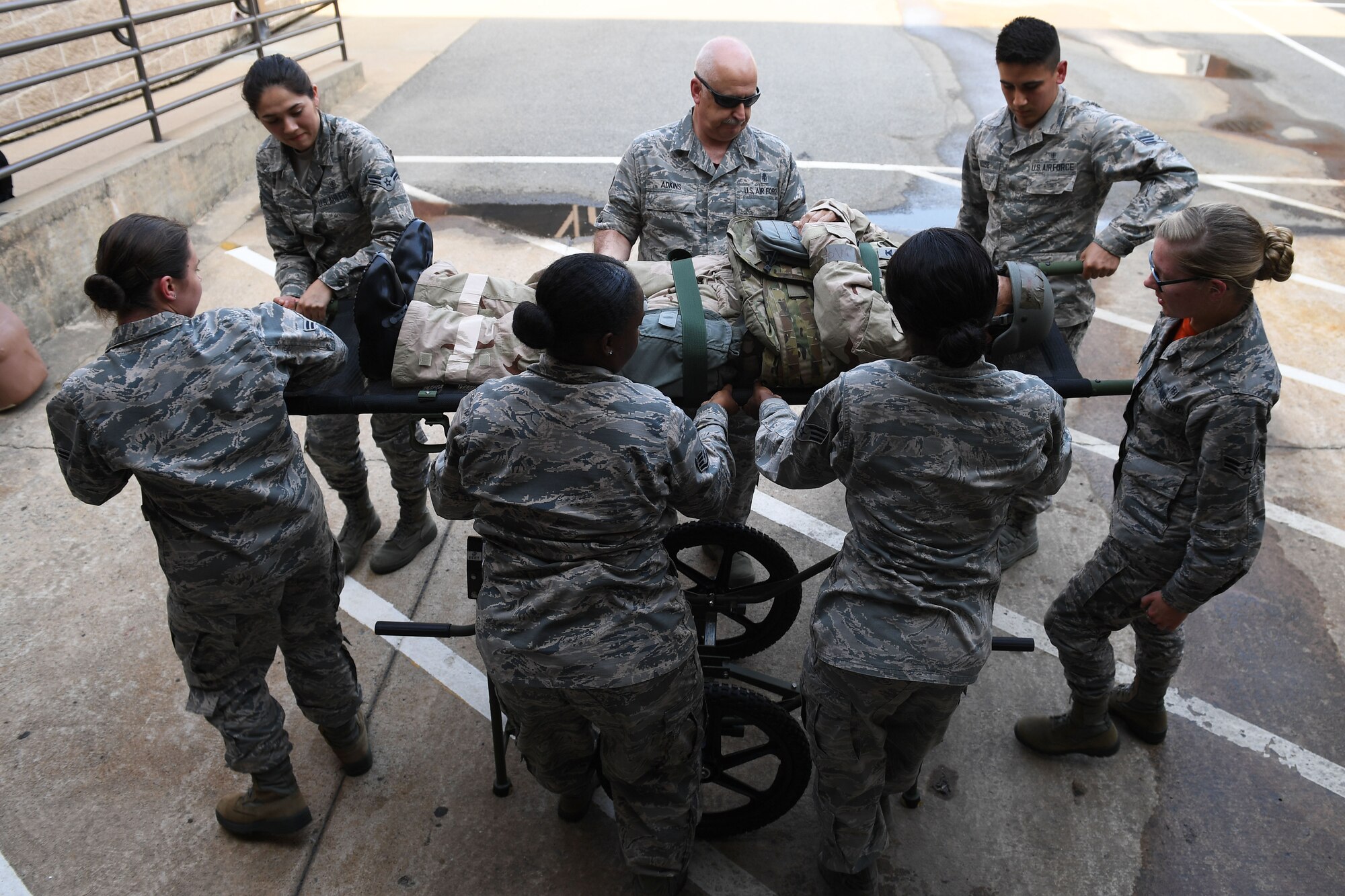 Seven green camouflaged Airmen gather around a medical gurney to practice strapping a patient onto it.