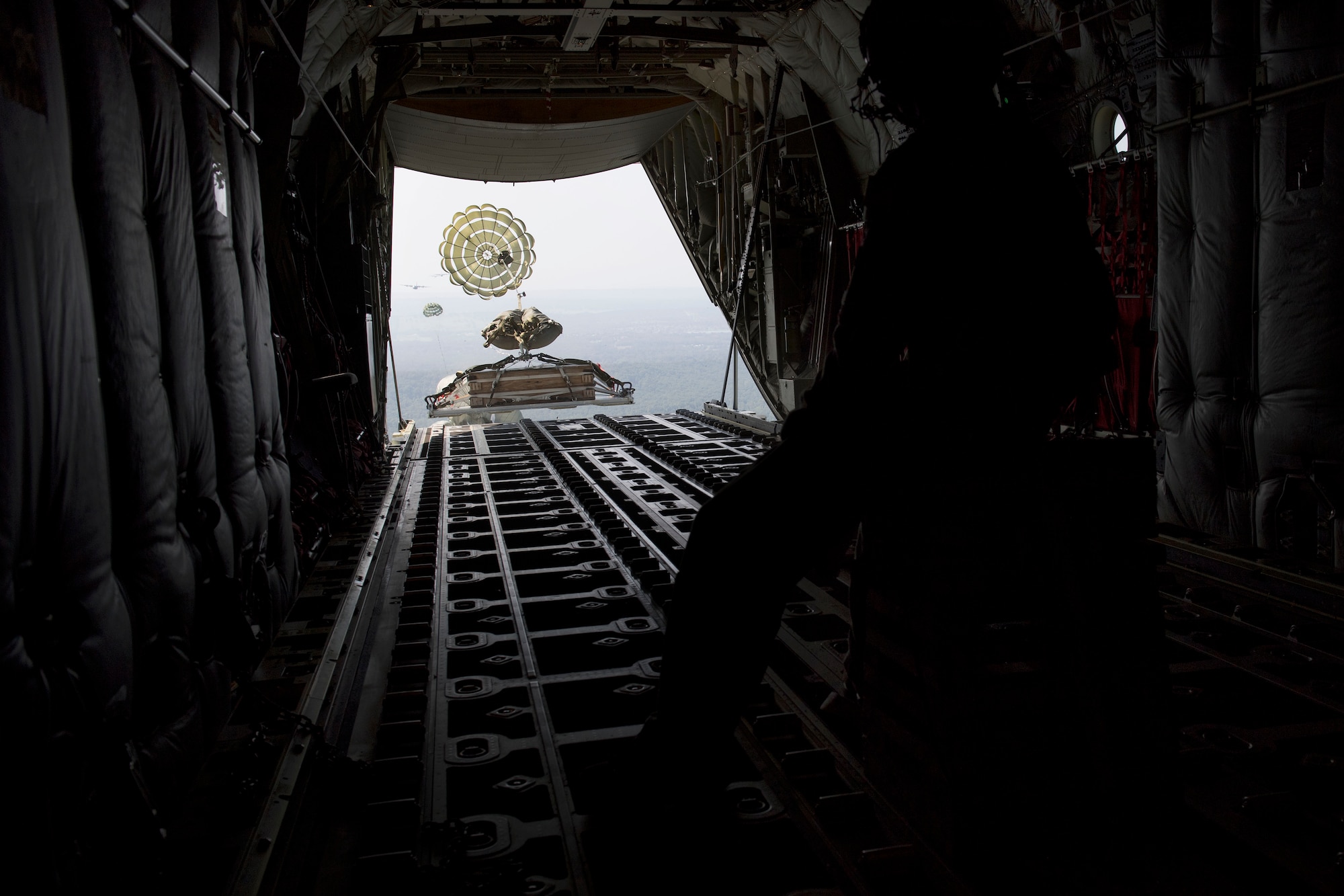 A long shot from the inside of a C-130J aircraft shows a parachuted pallet simulating heavy equipment sliding out the open back of the plane.