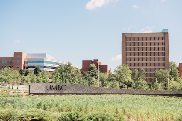 Landscape photo of UMBC sign at entrance to the campus during a sunny day. Three academic buildings are visible behind the sign.