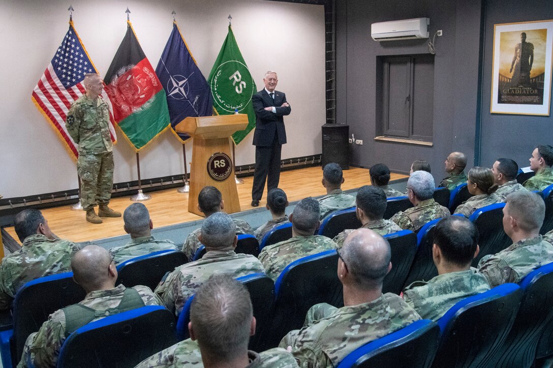 Defense leaders stand on a stage and talk to military officials in seats.