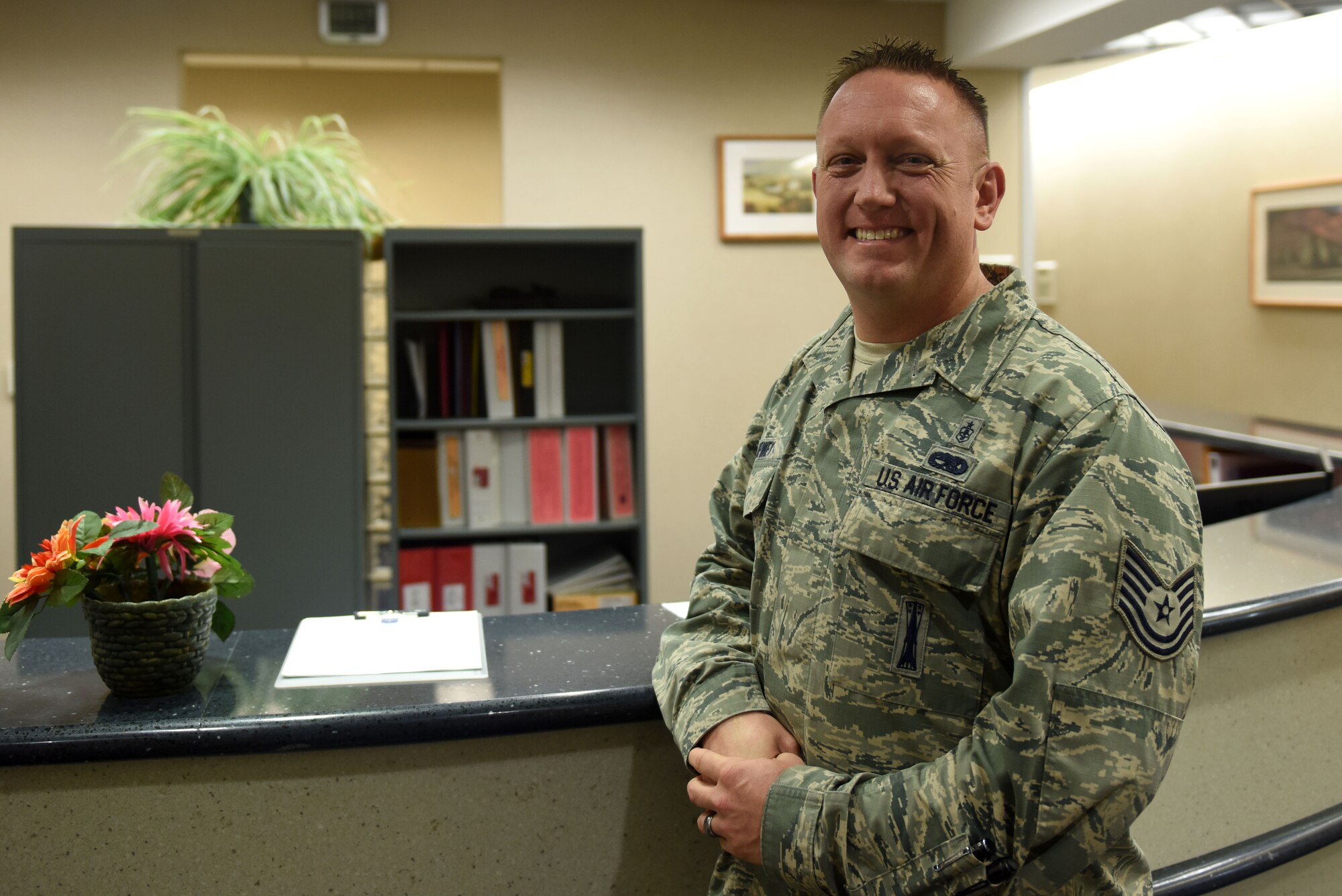 A man in the Airman Battle Uniform stands in front of a desk.
