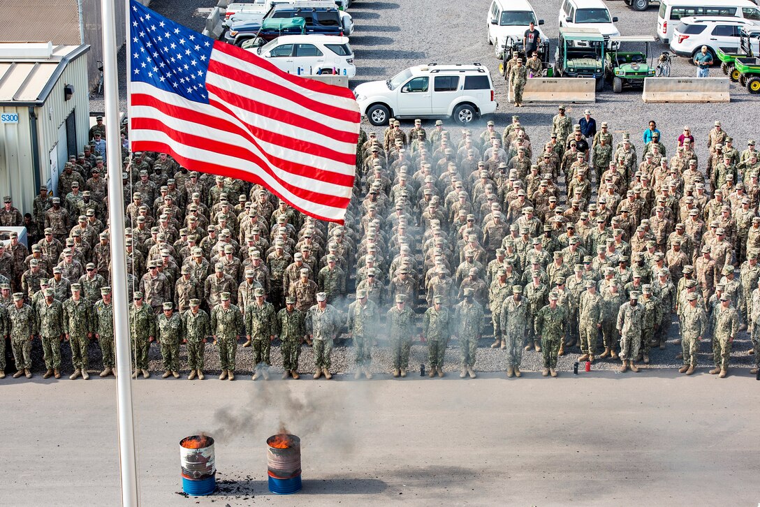 Service members stand at attention facing a U.S. flag and two burning barrels.