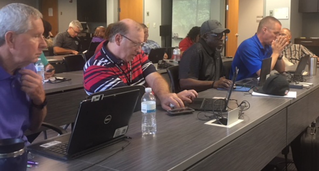 Group photo of various employees sitting in a room looking at their laptops.