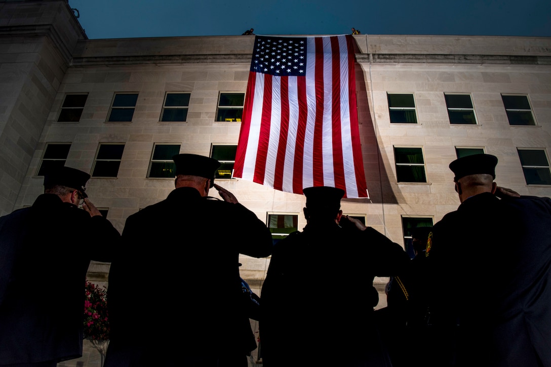 Four officers, shown from behind, salute a giant American flag on the side of the Pentagon.
