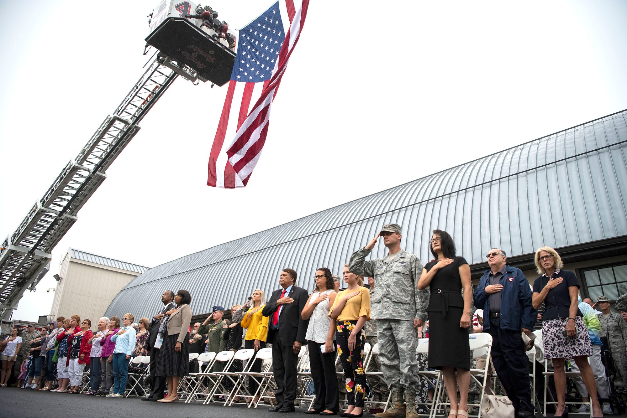 Attendees stand during the National Anthem at a 9/11 Memorial Service Sept. 11, 2018, at the Air Mobility Command Museum on Dover Air Force Base, Delaware. The large flag overhead was suspended from the ladders of firetrucks from Camden-Wyoming and Dover fire companies.