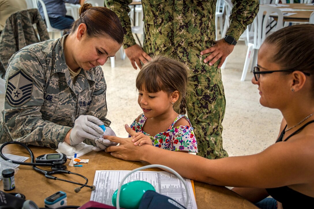 A service member holds a patient's hand while performing a medical procedure as a smiling girl watches.
