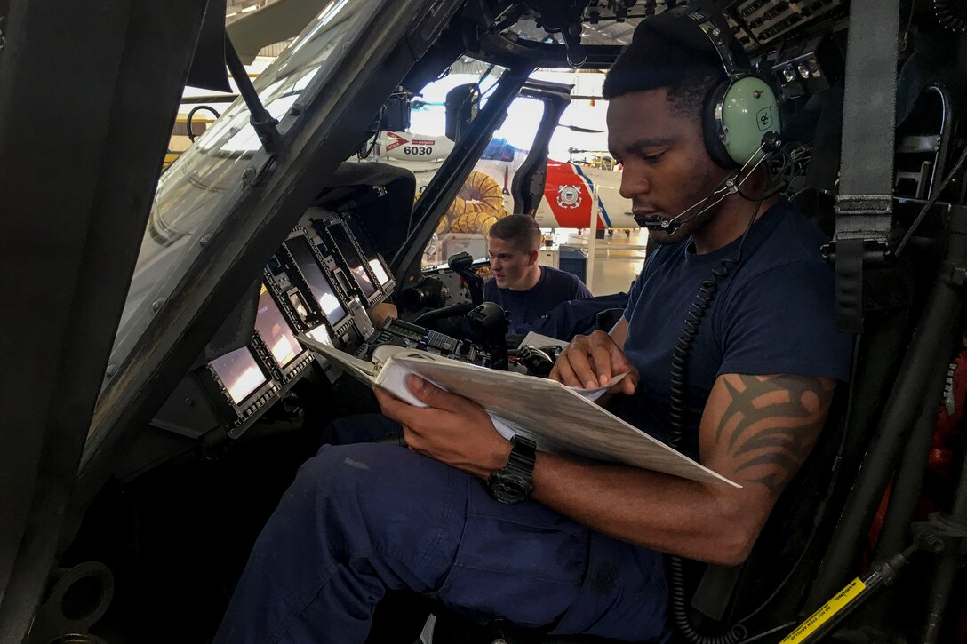 A Coast Guardsman reads a manual while another checks the instruments on a helicopter.