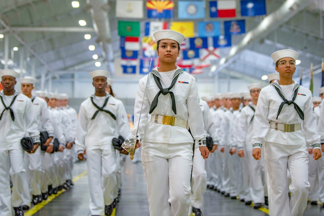 Sailors march in a large room with flags overhead.