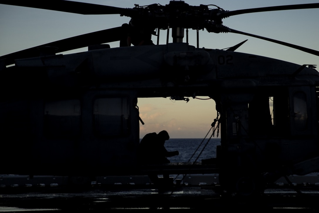 A Sailor with Helicopter Sea Combat Squadron 25 sits inside a U.S. Navy MH-60S Seahawk helicopter atop the flight deck of the amphibious assault ship USS Wasp (LHD 1) as the sun sets off the coast of Okinawa, Japan, Aug. 25, 2018.
