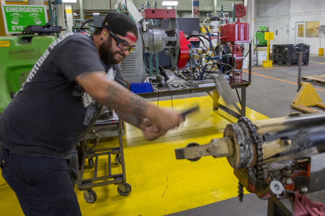 Cory Moya, heavy mobile equipment mechanic, uses a sledge hammer to dislodge a seized hydraulic cylinder at the production plant aboard Marine Corps Logistics Base Barstow, Calif. Moya explains that the reservoir had a leak and may need to be repaired for quality control purposes.