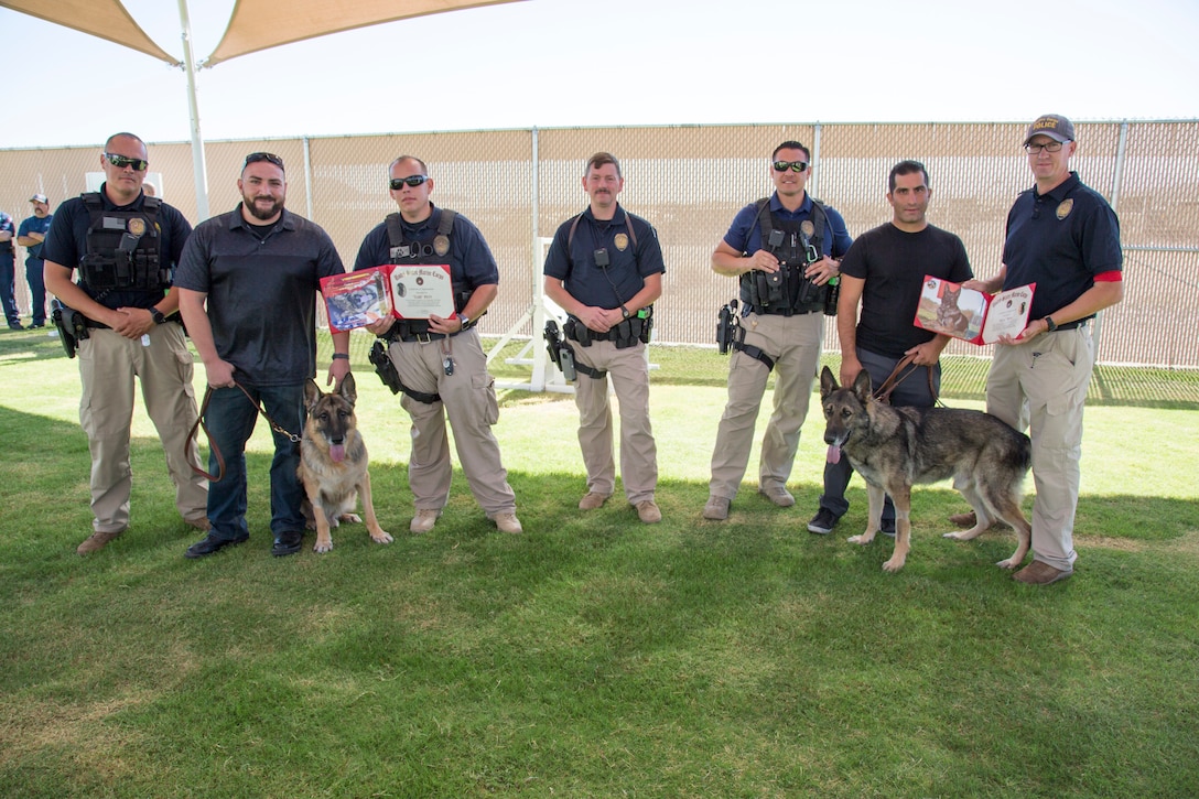 Jacob Lucero(second for left) and Nadeem Seirafi ((second from right) hold the leashes of Colli and Ricsi during the two Military Workings Dogs' retirement ceremony at the K-9 Training Field adjacent to the Adam Leigh Cann Canine Facility aboard Marine Corps Logistics Base Barstow, Calif., Sept. 12. The two handlers partnered with their K-9 companions on the beat and are now adopting the two dogs as they retire from active duty. Lieutenant Steven Goss, (far right) kennel master, Marine Corps Police Department, presented the two dogs with retirement certificates. The other officers in the photo also partnered with Colli and Ricsi at different times in their careers.