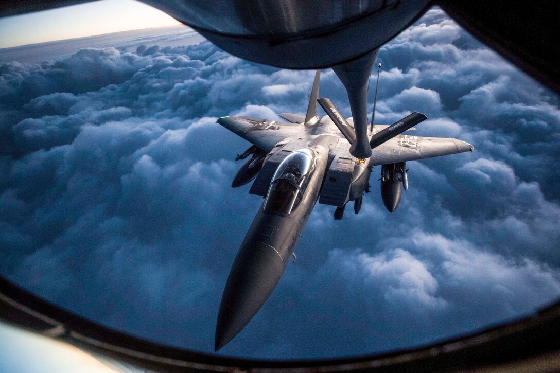 An aircraft receives fuel from the boom of another aircraft while flying in blue clouds.