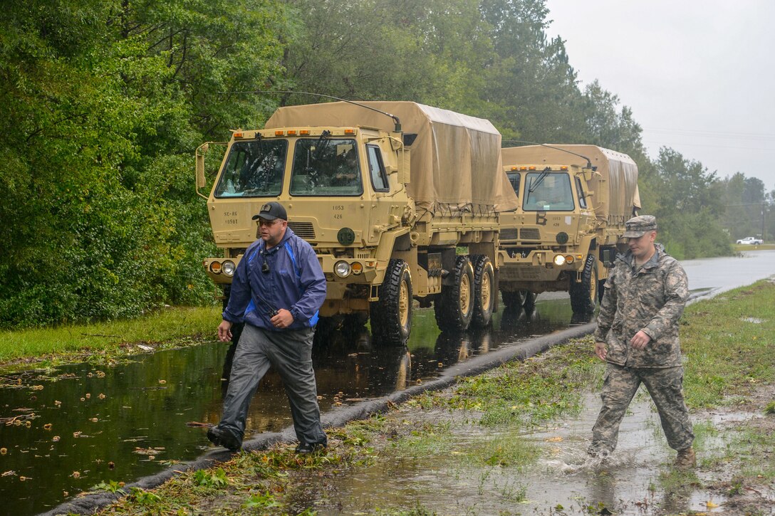 Two men walk through a flooded area while two military trucks sit on the road in the background.