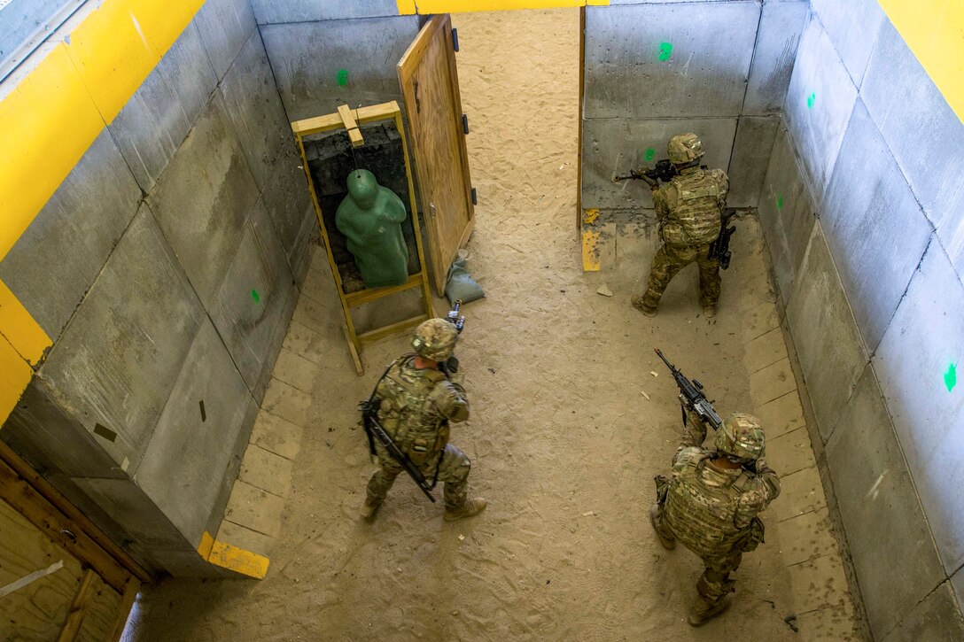 Three soldiers, shown from overhead, aim weapons in a structure with cement block-type walls and a dirt floor.
