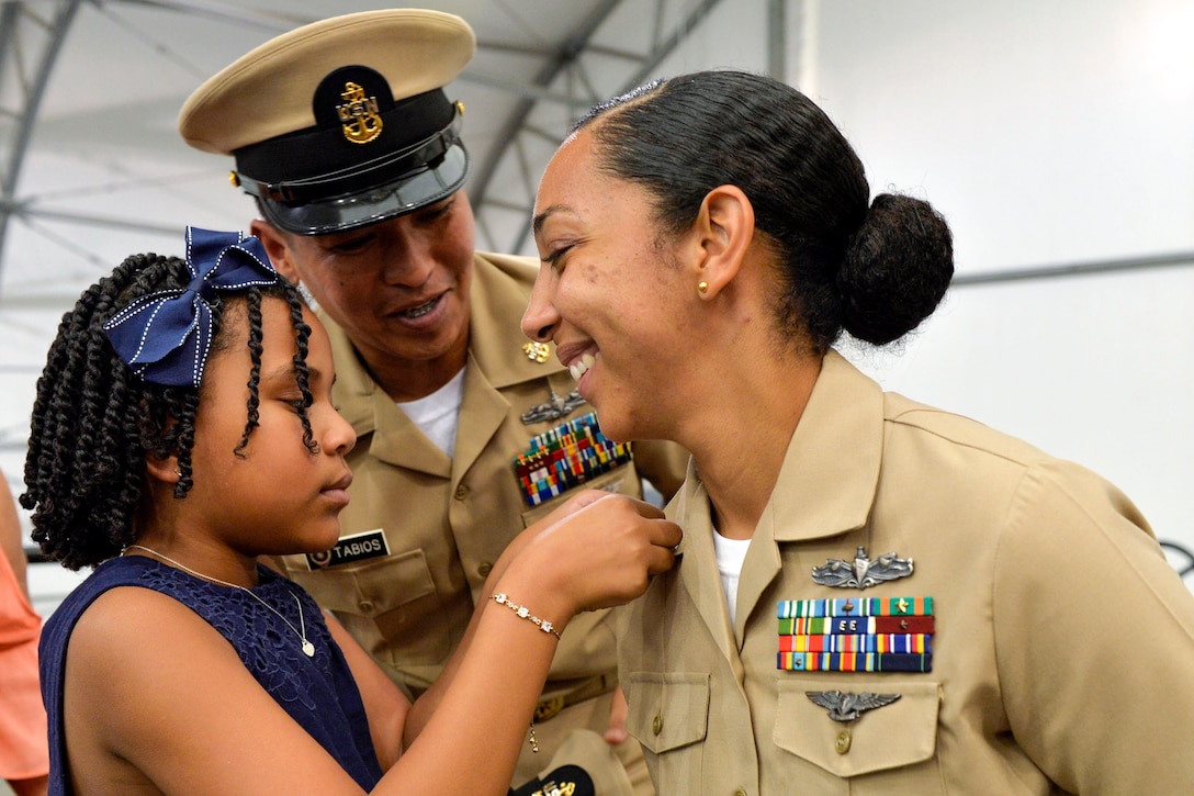 A child affixes a pin to the lapel of a smiling sailor as another sailor watches.