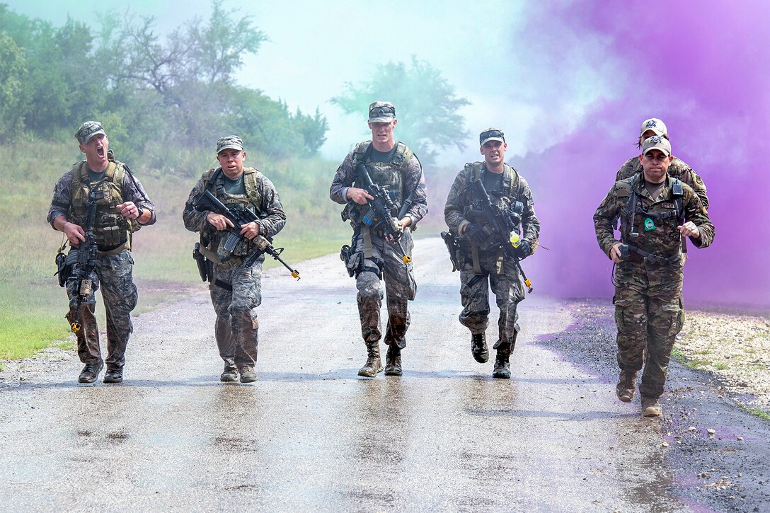 Six airmen carrying weapons jog on a road with purple smoke in the background.