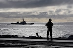 A sailor stands on the flight deck of the aircraft carrier USS Harry S. Truman during flight operations in the North Atlantic as the guided missile cruiser USS Normandy sails alongside.