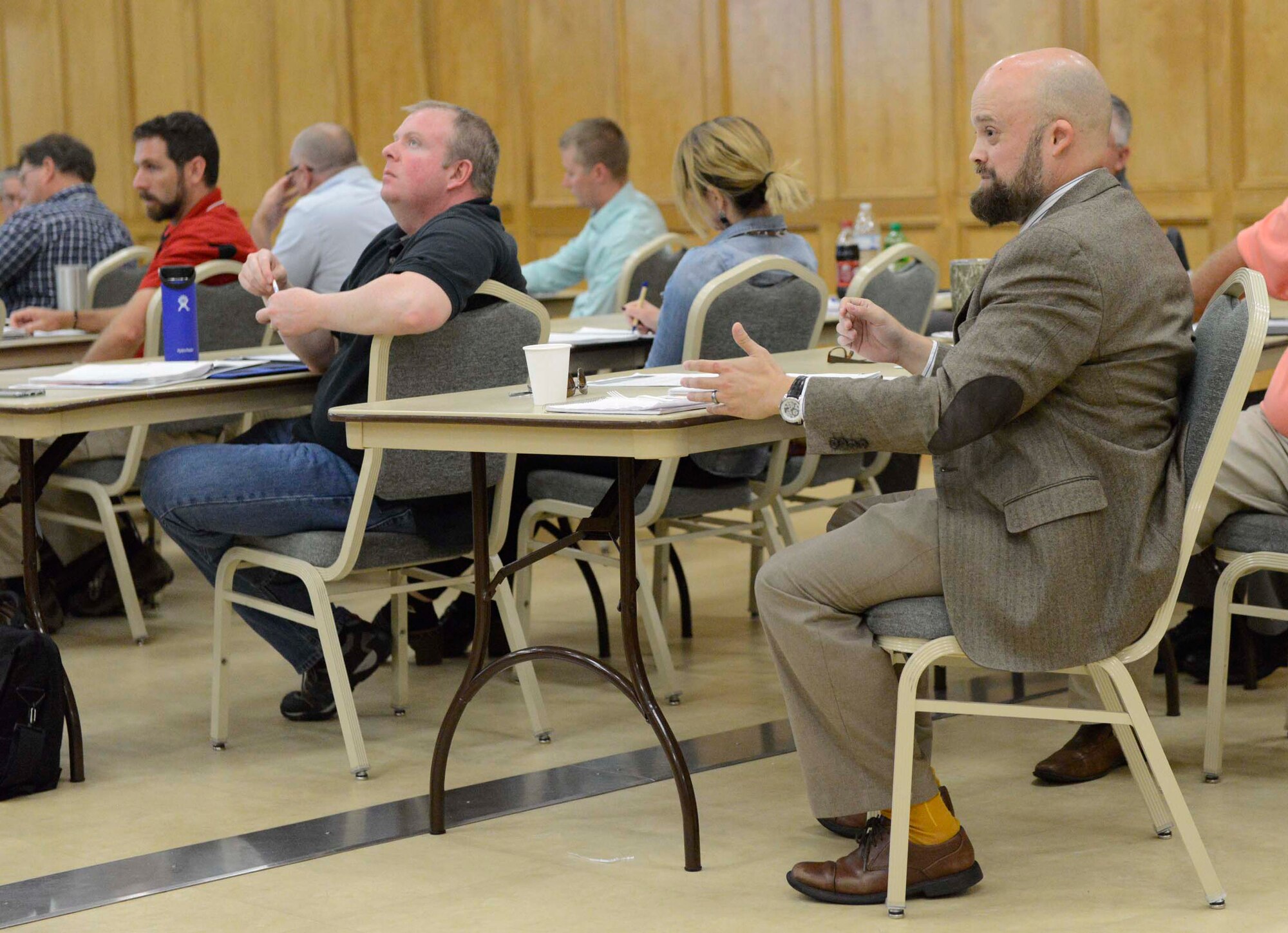 A bald, bearded white male in brown suit sits at a table in a room full of people sitting at tables.