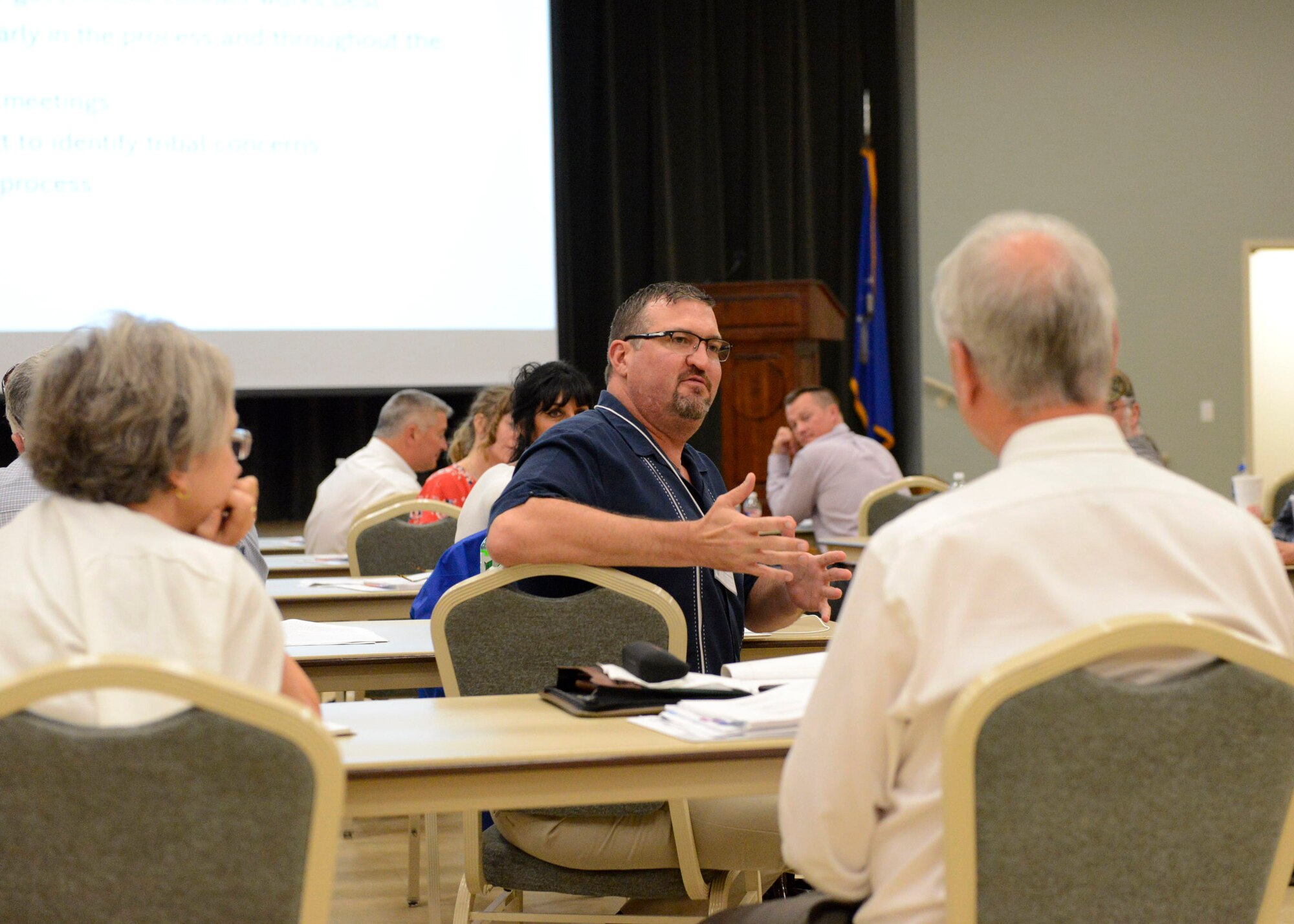 A dark-haired, bearded white male sits at a table and talks to other people talking at tables.