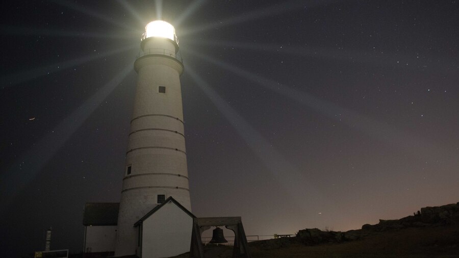 Beams of light emanate out on a dark gray sky from a white lighthouse.