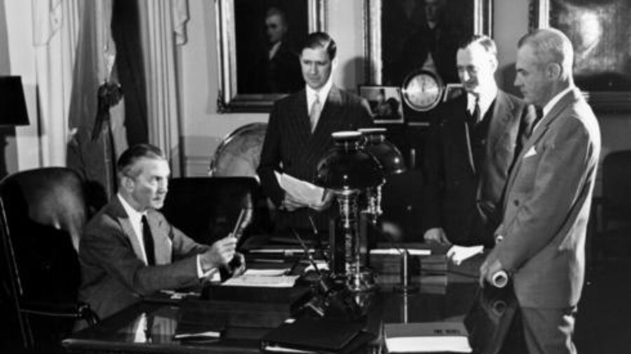 Three men stand around a desk where another man is seated.