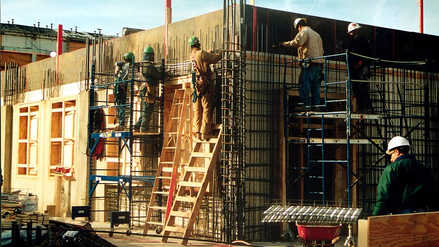 Construction personnel on ladders and scaffolding work with steel rods against a wood facade.