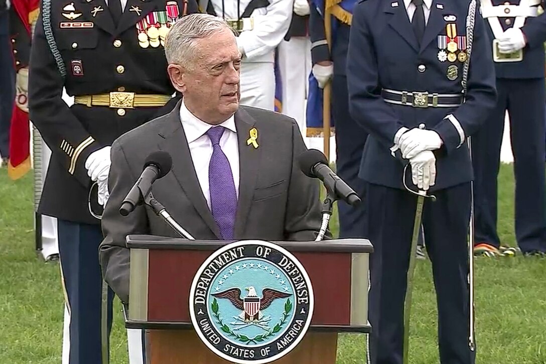 Defense Secretary James N. Mattis speaks at a lectern on a parade field, as troops stand behind him.