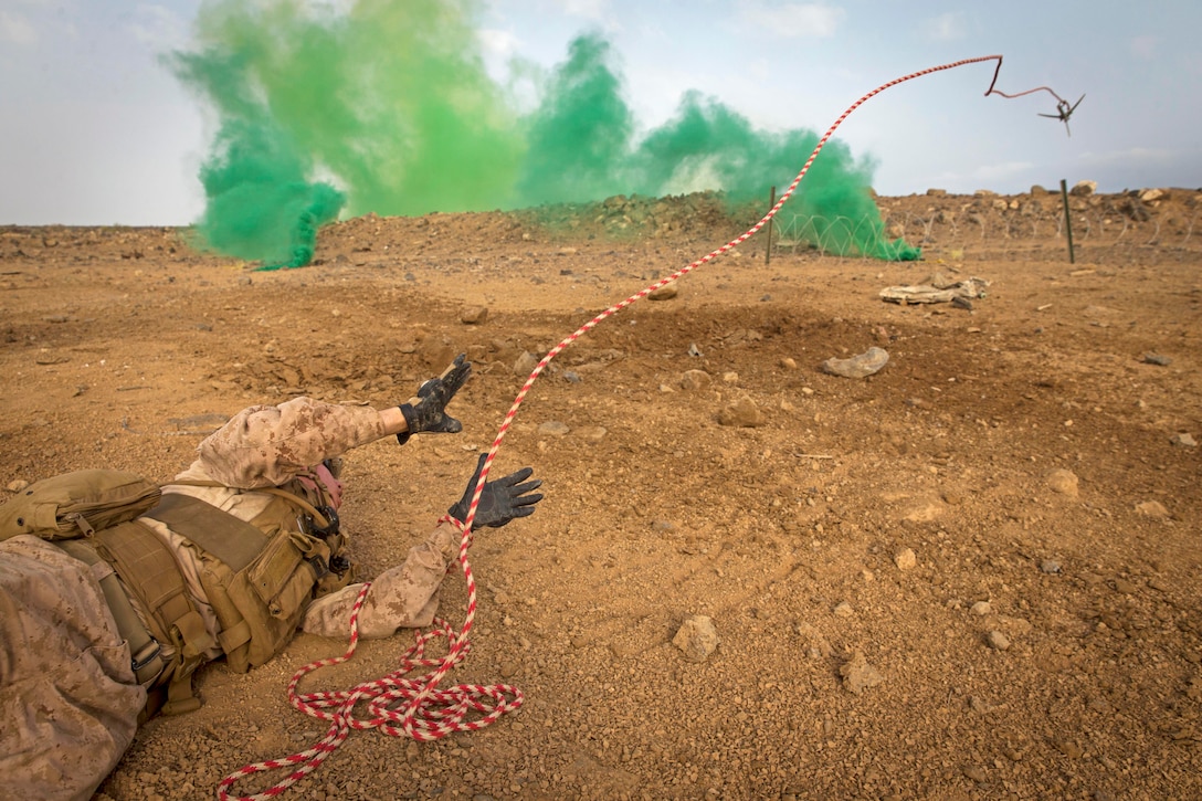 A Marine throws a hook from his position on the ground with green smoke in the foreground.