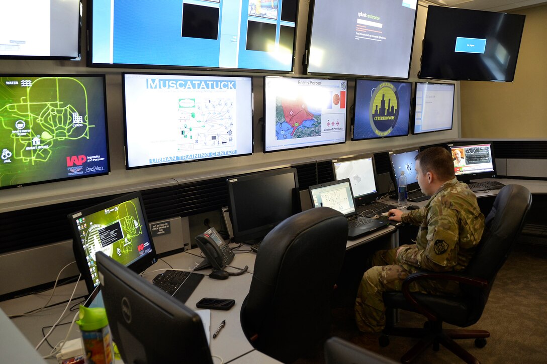 An Army officer sits in front of a bank of computer monitors.