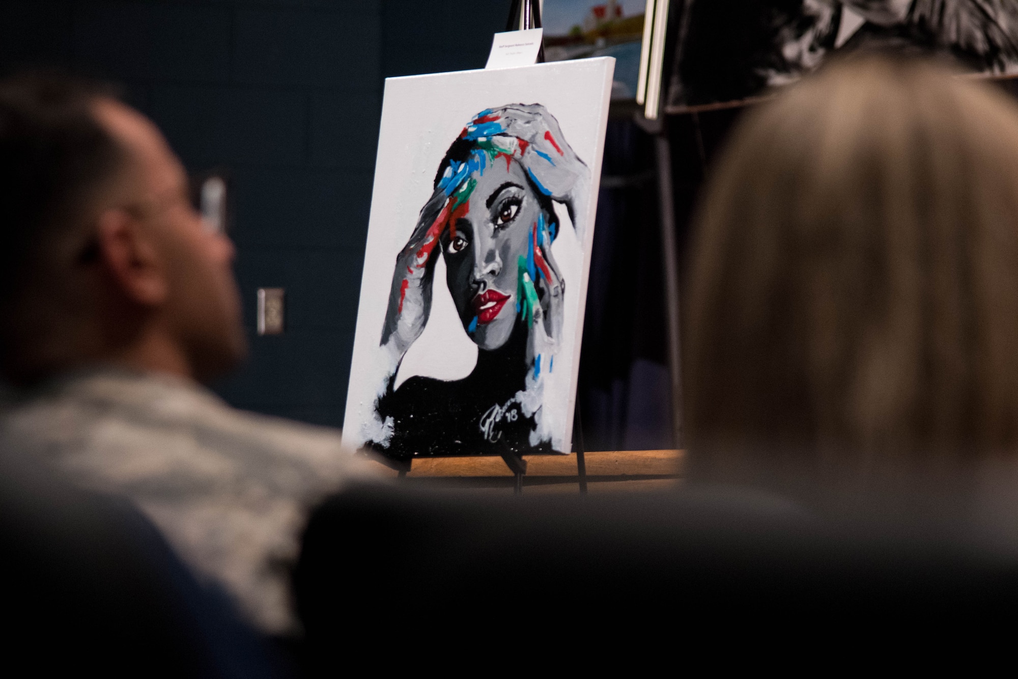 The painting “Healing Touch” created by U.S. Air Force Staff Sgt. Rebecca Salcedo, Air Combat Command Public Affairs, is seen between two audience members at the “Creating Change Through Art” event held at the Langley Air Force Base Theater, Virginia, Sept. 25, 2018. The painting was part of an art contest promoting healthy outlets that allow service members to cope with difficult emotions. (U.S. Air Force Photo by: Senior Airman Ericha Guyote/Released)