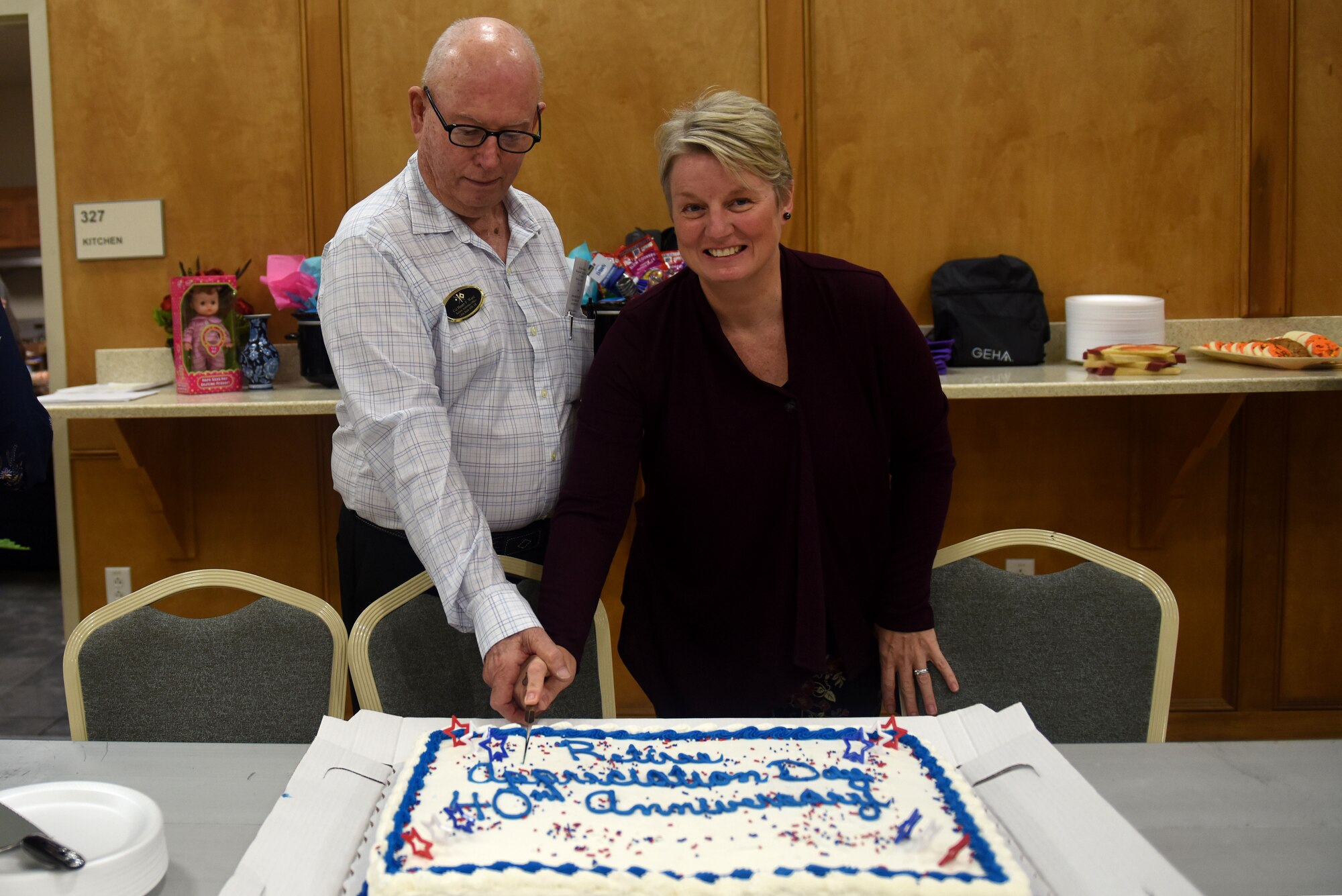 A man in a white button up shirt and a woman in a black blouse cut a cake with white and blue frosting.